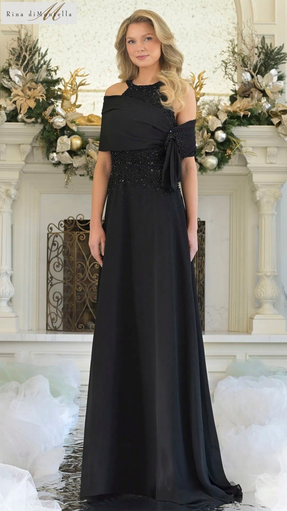 A woman stands indoors by a decorated fireplace, wearing the Rina Di Montella RD3304 long formal dress with a black beaded bodice and flowing chiffon A-line skirt.